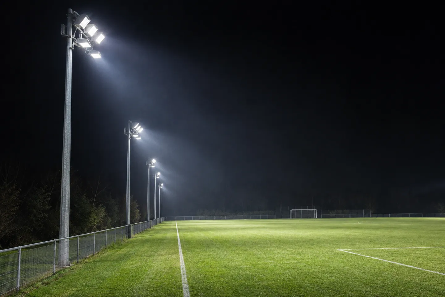Pali con proiettori a LED posizionati ai lati di un campo da calcio amatoriale e puntati verso il campo di notte, a dimostrazione di come l’altezza e la disposizione dei pali influenzino la progettazione dell’illuminazione sportiva.