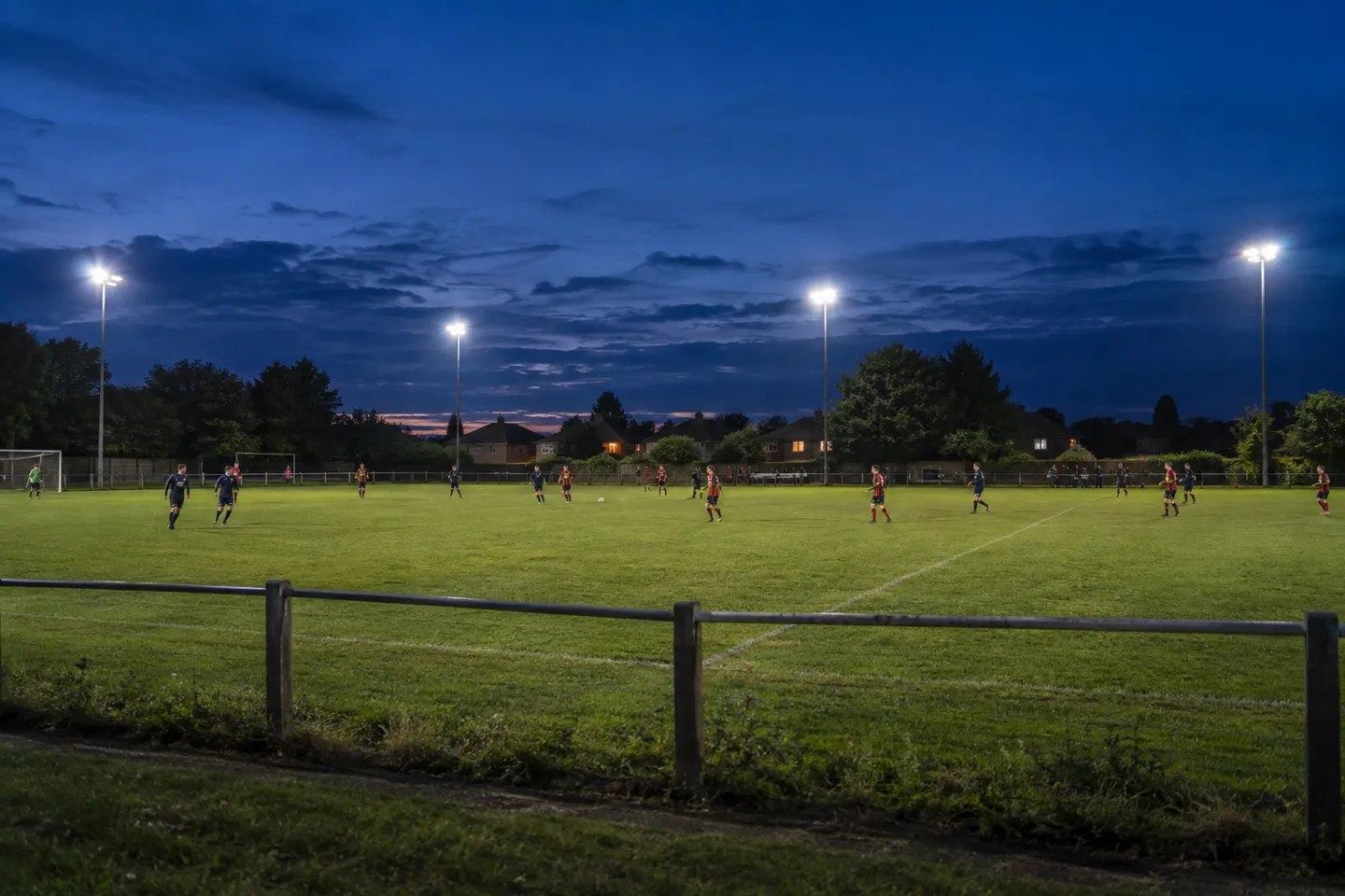 An amateur community football match under four modest floodlight poles at dusk — the low end of the stadium lighting cost and running-cost scale.